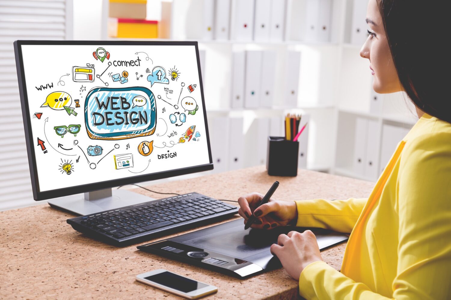 A young woman in a yellow shirt draws a colorful web design sketch on her desktop computer.