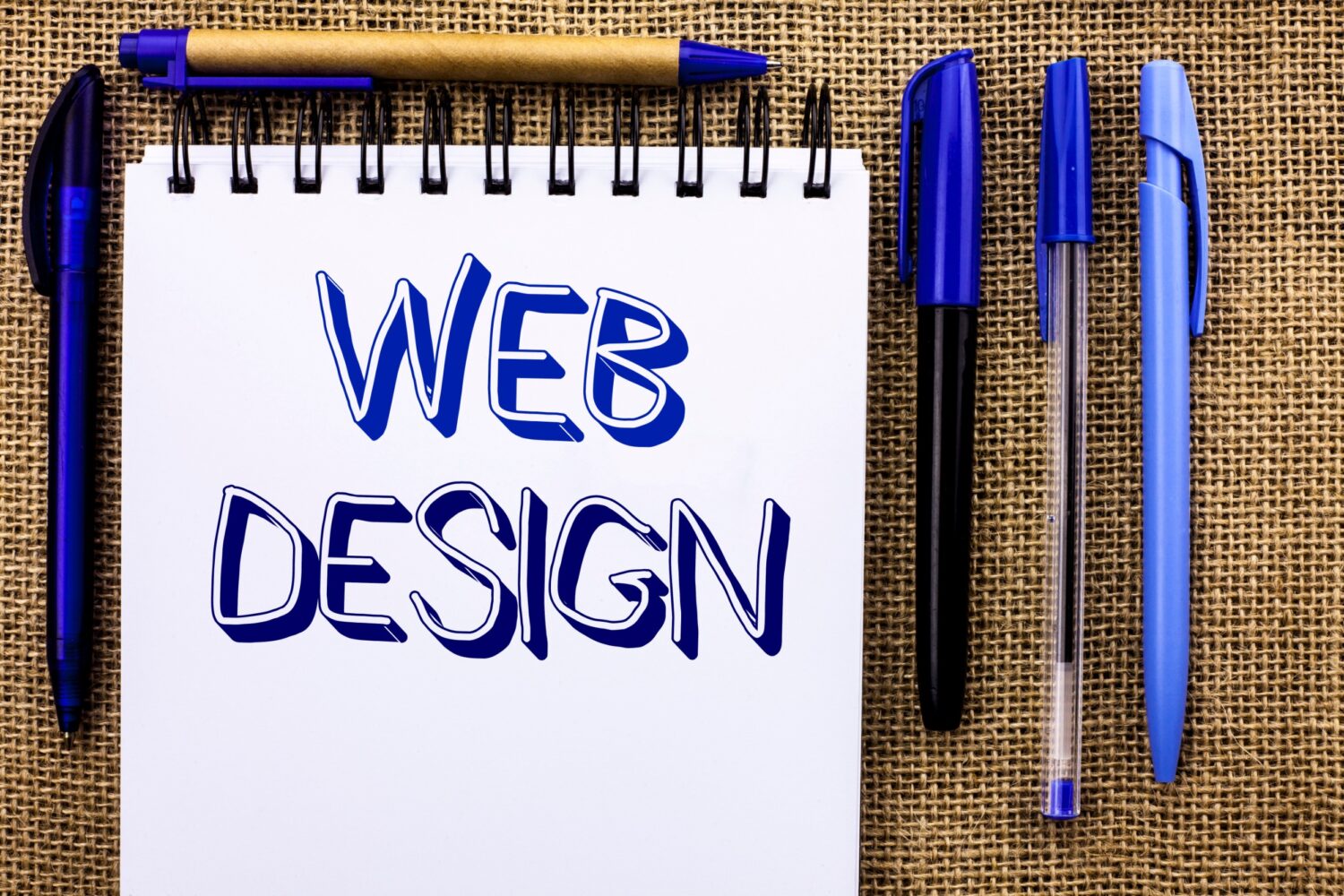 A notebook with the words “Web Design” written across the page is surrounded by pens.